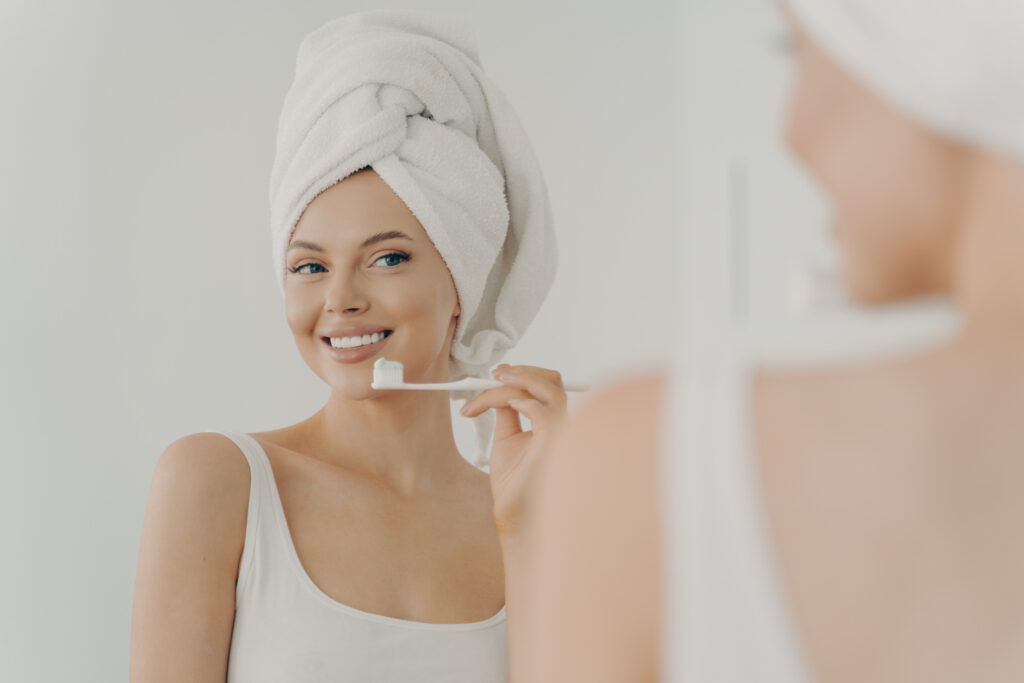 Junge Frau hält Zahnbürste in der Hand und blickt lächelnd in den Spiegel
