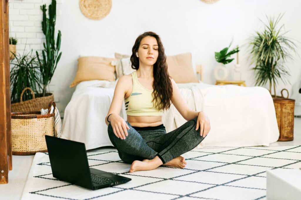 Frau in Trainingsgewand sitzt am Teppich vor Laptop und meditiert