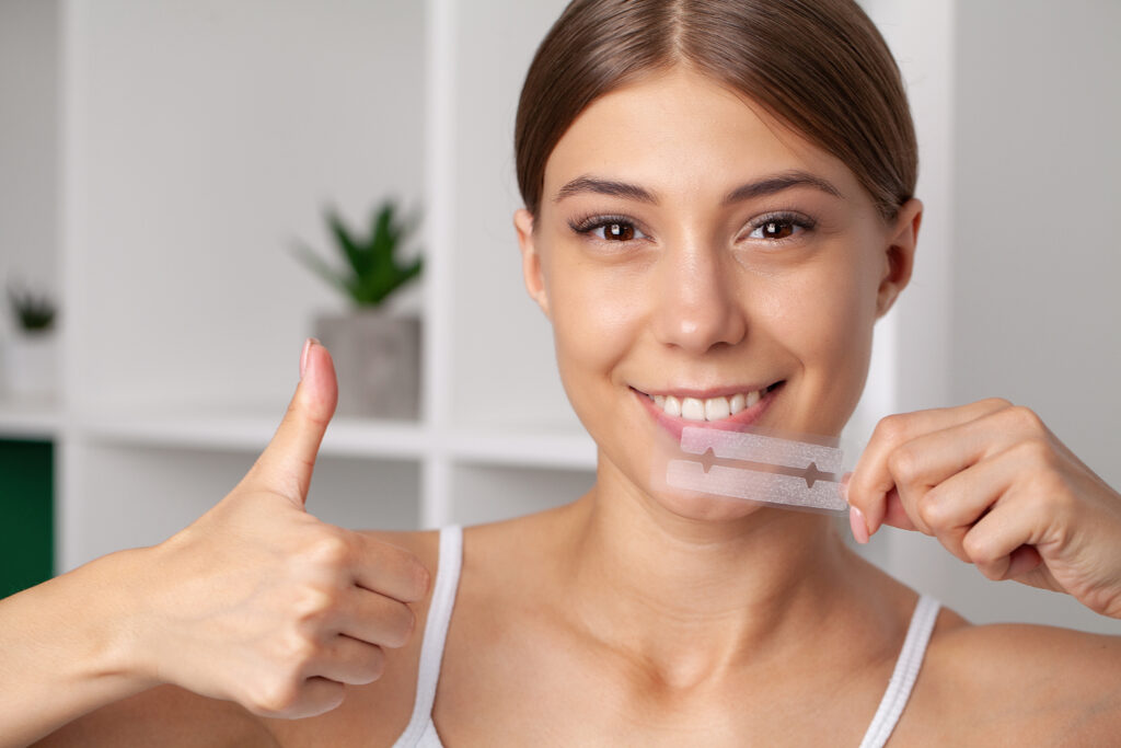 Lächelnde Frau hält Zahnaufhellungsstreifen in der Hand und zeigt den Daumen