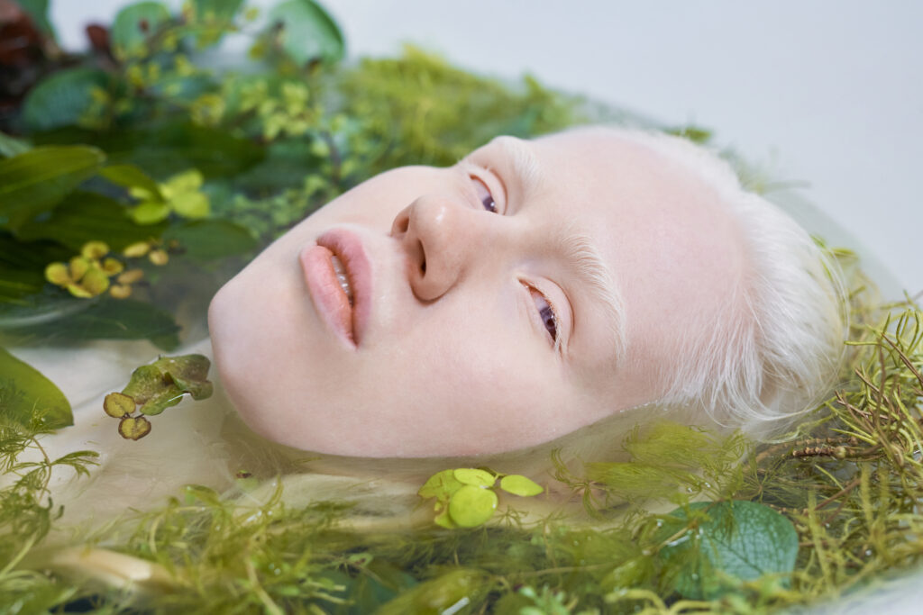 Junger Mann mit blonden Haaren und blonden Augenbrauen liegt in einem Becken mit Blättern