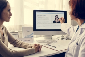 Zwei Damen in heller Kleidung sitzen vor einem PC. Die eine Dame zeigt mit einem Stift und ein Bild auf dem PC.