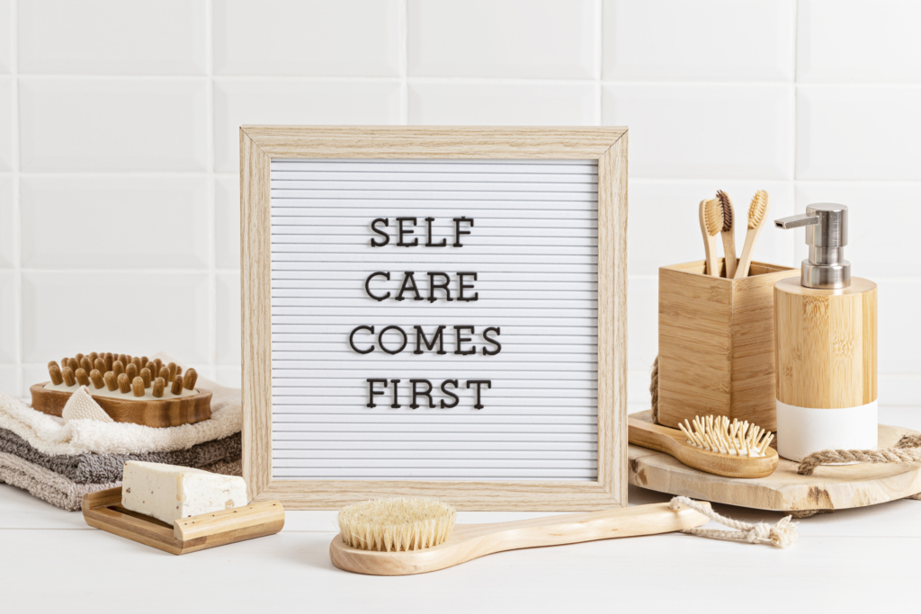 Schild mit Aufschrift: Selfcare comes first. Daneben rechts ein Seifenspender, eine Bürste und ein Holzbecher mit Zahnbürsten; links drei kleine Handtücher und eine Bürste