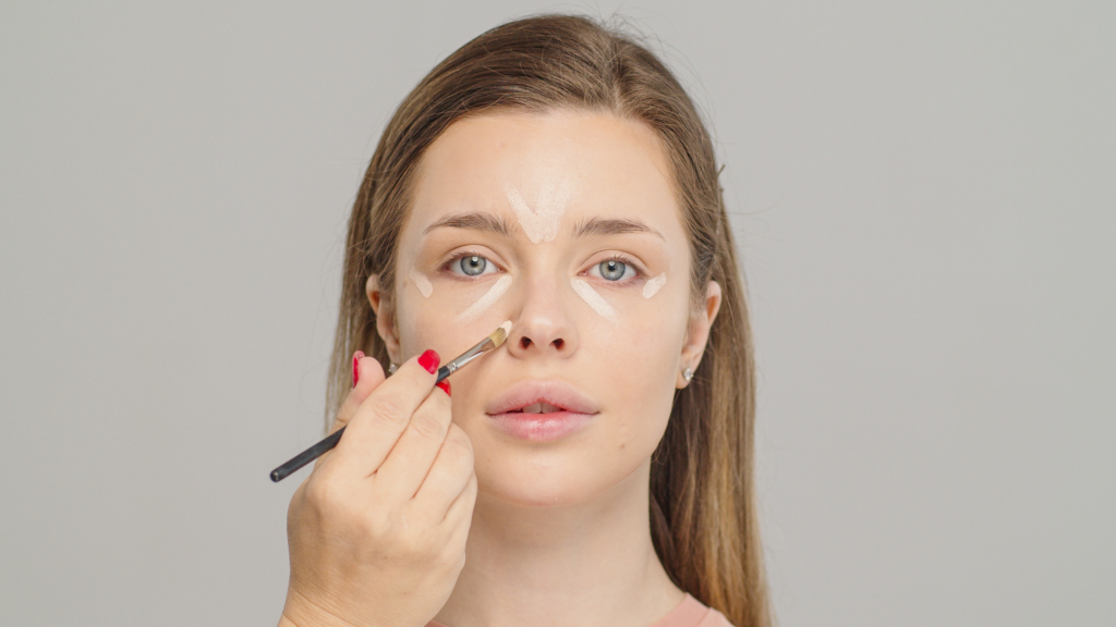 Junge Frau mit langen braunen Haaren, die ein paar Concealer-Striche in ihrem Gesicht hat. Ein Pinsel wird von einer Hand mit roten Fingernägeln geführt