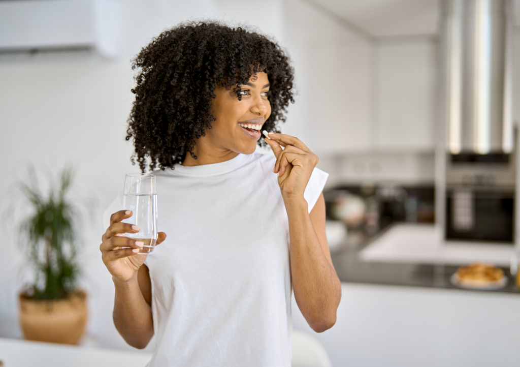 Frau mit dunklen Haaren und weißém T-Shirt steht in einer Küche. Sie hält ein Glas Wasser in einer Hand und eine Kapsel, die sie zum Mund führt, in der anderen