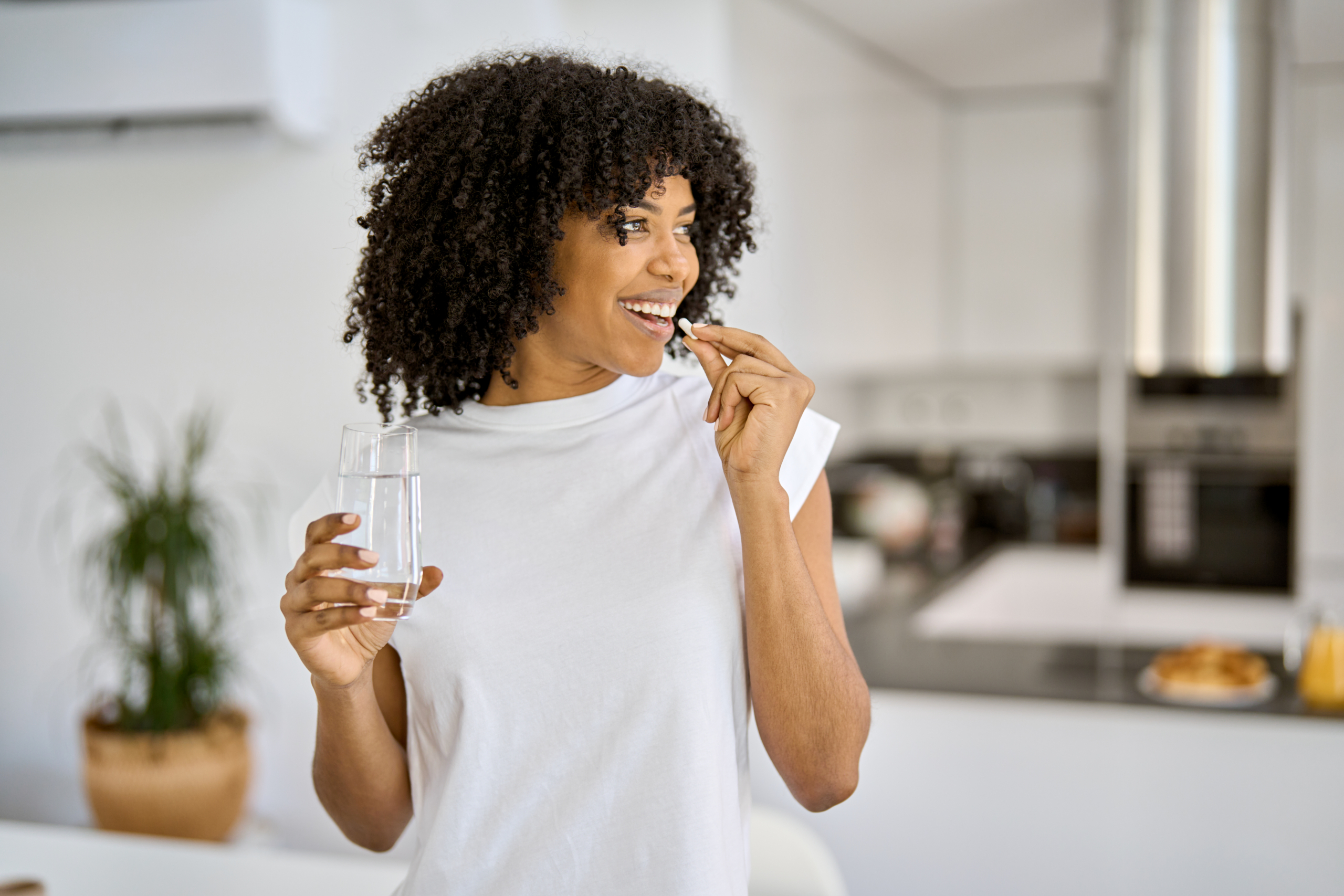 Frau mit dunklen Haaren und weißém T-Shirt steht in einer Küche. Sie hält ein Glas Wasser in einer Hand und eine Kapsel, die sie zum Mund führt, in der anderen