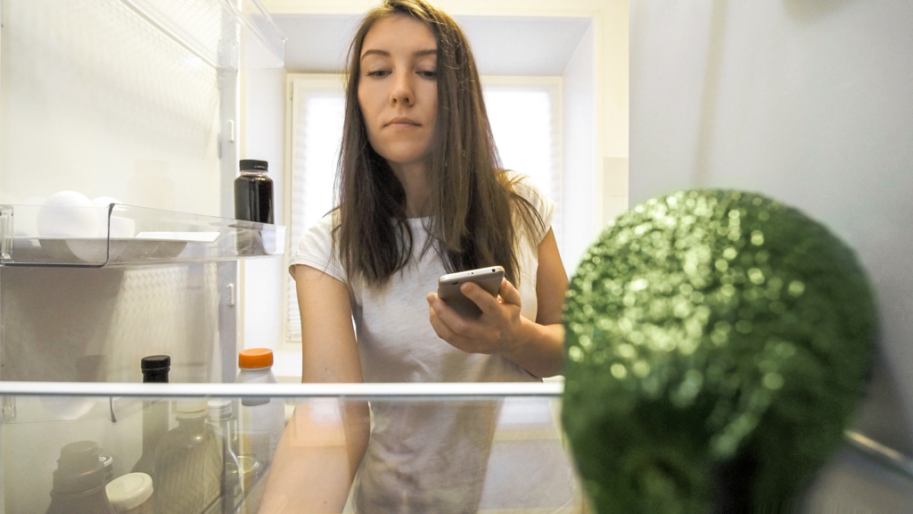 Frau hält Handy in der Hand und blickt in den Kühlschrank