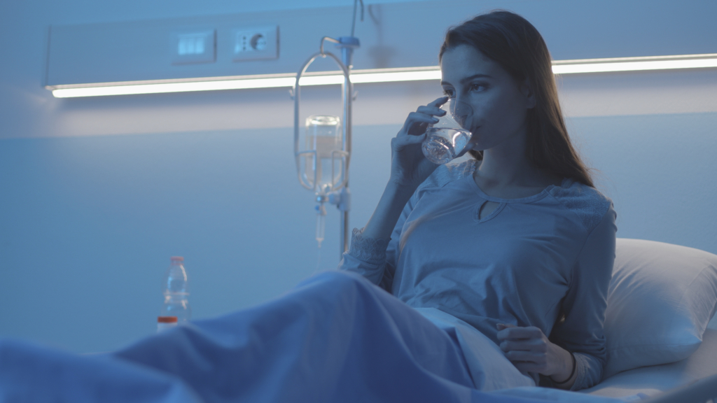Frau die in einem Krankenhausbett liegt und für die Post-OP Pflege ein Glas Wasser trinkt