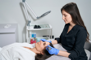 Frau liegt auf einer Kosmetikliege. Hinter ihr steht eine andere Frau mit schwarzem Kleid und blauen Hygienehandschuhen und hält einen Laser auf ihre Stirn