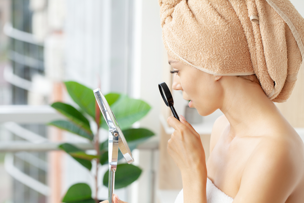 Frau mit einem Handtuch über die Haare gewickelt hält eine Lupe in der Hand und schaut in einen Kosmetikspiegel