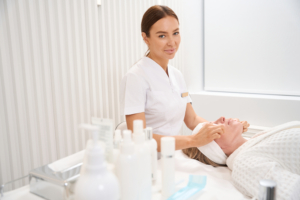 Kosmetikerin in weiß gekleidet steht hinter einer Kundin auf einer Kosmetikliege und hält ihr Gesicht