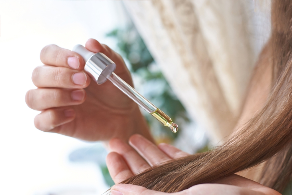 Hand hält eine Pipette mit Haaröl. Die andere Hand hält eine Haarsträhne