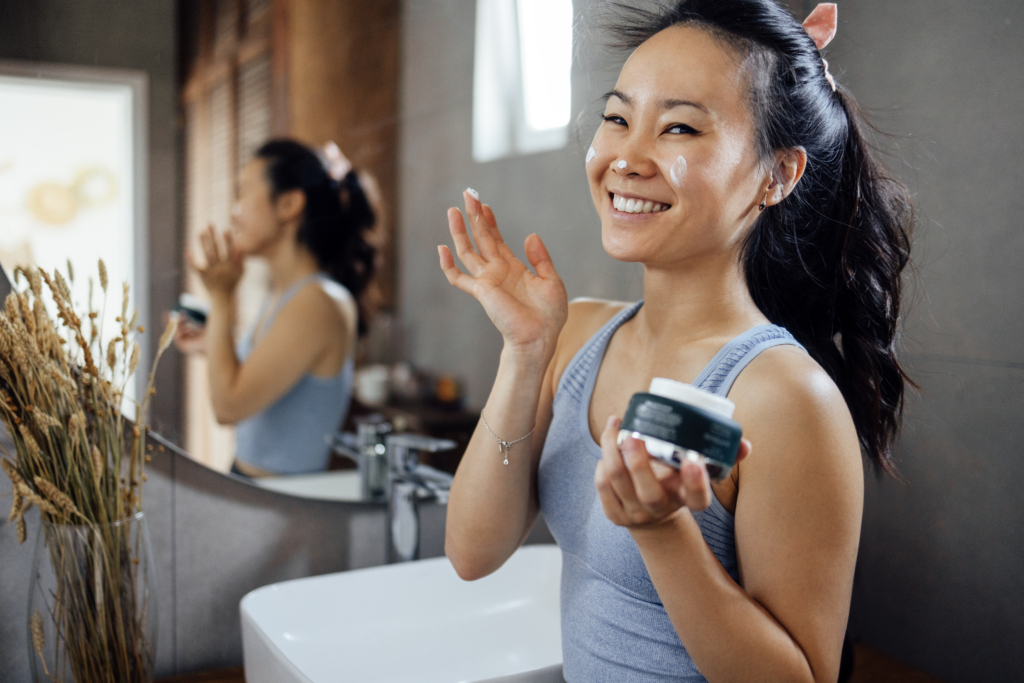 junge Frau die Creme in ihr Gesicht gibt, dabei Lacht und die Creme auch in der Dose in ihrer Hand hält